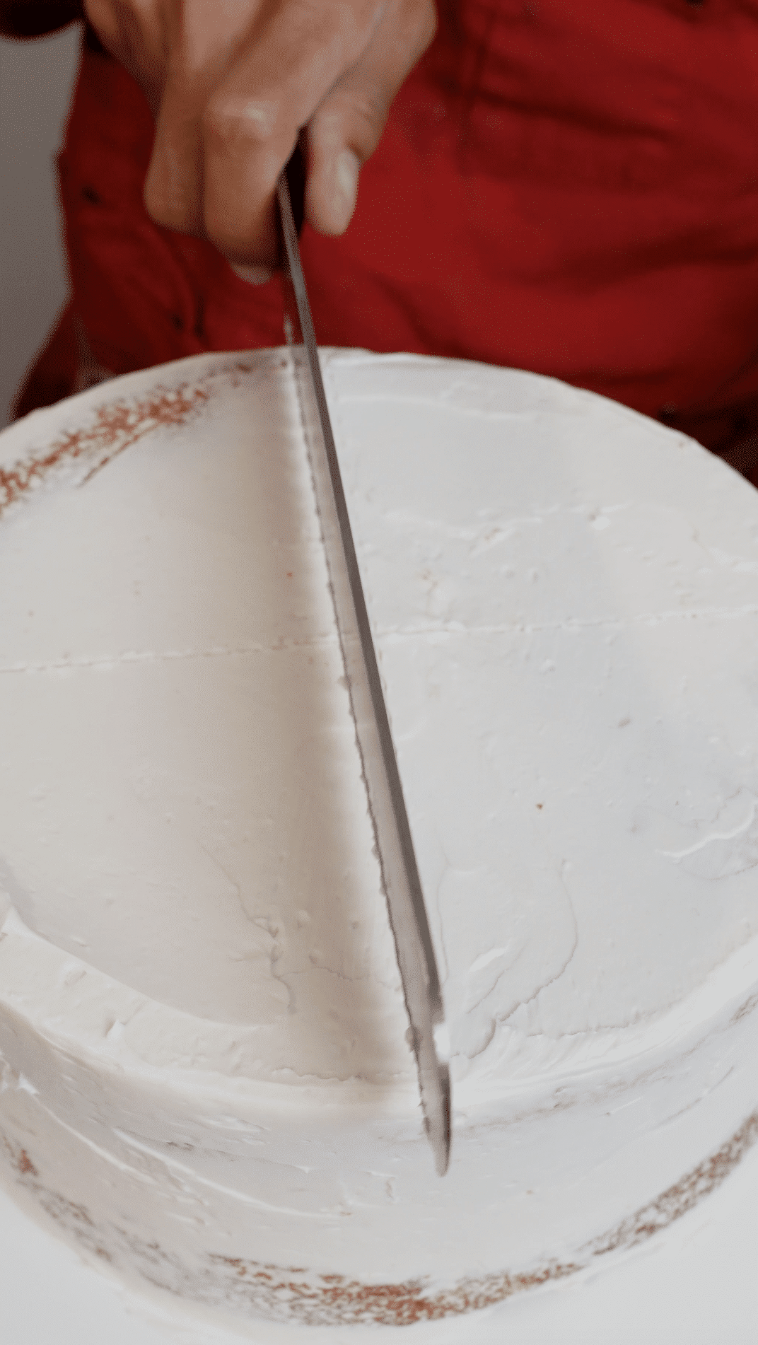 Baker using a serrated knife to lightly sketch a cross shape on top of a round cake to guide carving for a heart cake.